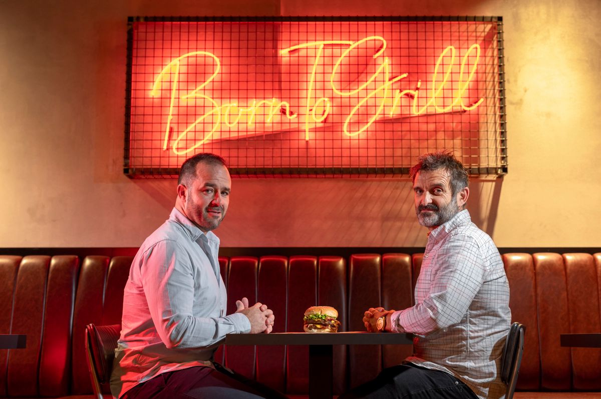 Adrian Stadelmann et Richard Williams assis à une table avec un hamburger au centre, devant un panneau lumineux 'Born to Grill' à Lausanne, Suisse.