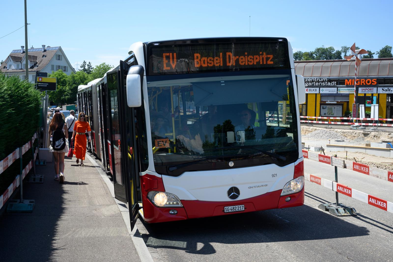 Gelenkbus mit Zielanzeige «EV Basel Dreispitz» an einer Haltestelle, im Hintergrund eine Migros-Filiale und Passanten. Gelenkbus mit Zielanzeige «EV Basel Dreispitz» an einer Haltestelle, im Hintergrund eine Migros-Filiale und Passanten.