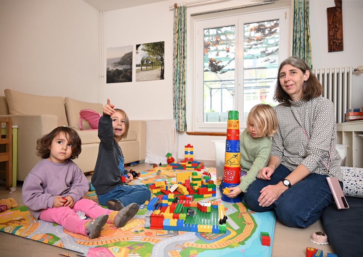 Tagesmutter Ursula Sylvestre sitzt mit drei Kindern auf einem Spielteppich. Die Kinder spielen mit bunten Bauklötzen im Wohnzimmer.