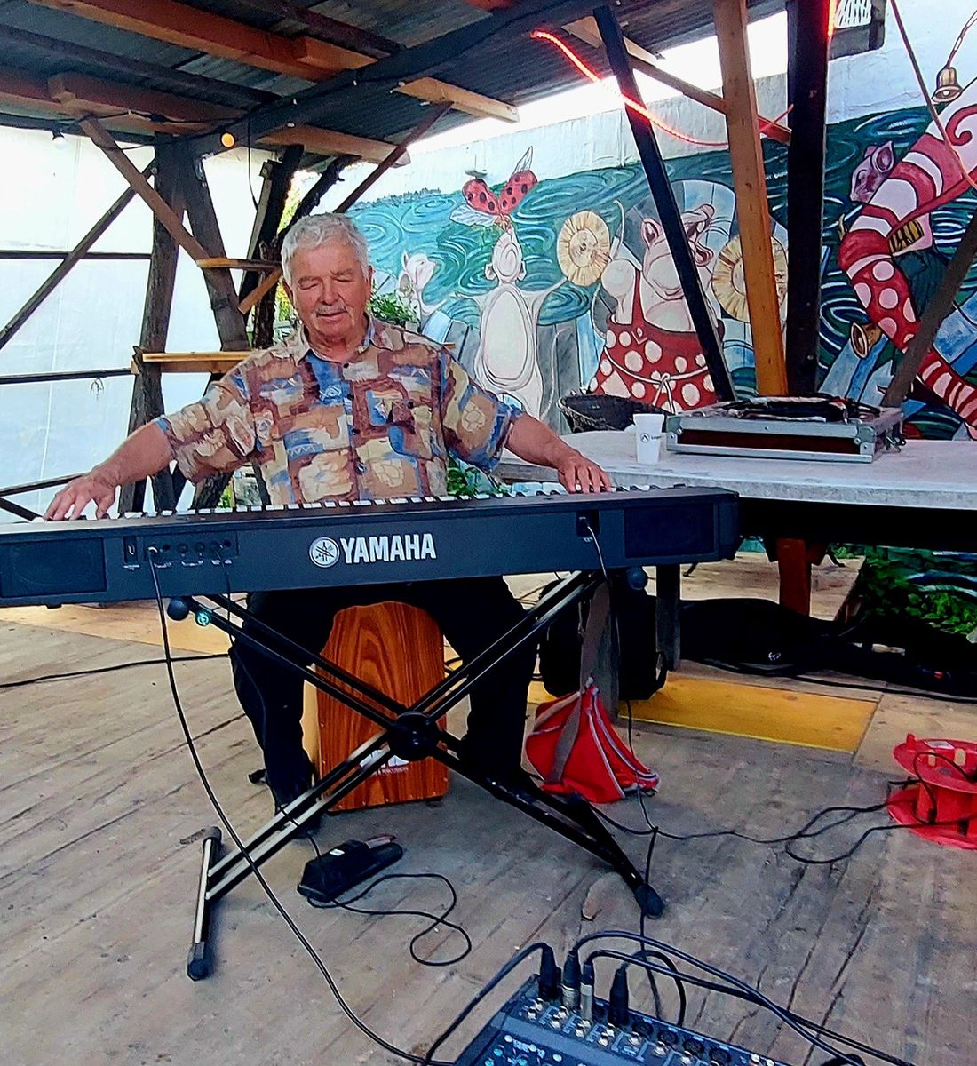 Un homme assis joue du clavier Yamaha sur une scène en bois, avec une peinture murale colorée en arrière-plan.