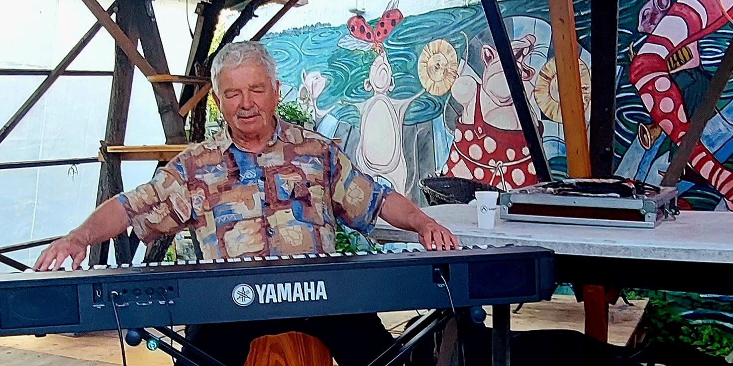 Un homme assis joue du clavier Yamaha sur une scène en bois, avec une peinture murale colorée en arrière-plan.