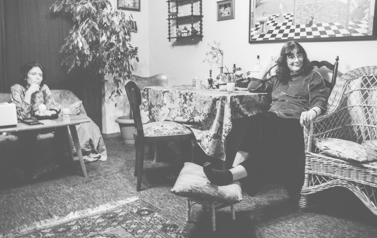 Adelheid Duvanel sitzt auf einem geflochtenen Sessel in einem Wohnzimmer, ihr Fuss ruht auf einem kleinen Hocker. Ihre Tochter sitzt im Hintergrund. Der Raum ist mit Teppichen und einem grossen Pflanzenarrangement dekoriert.