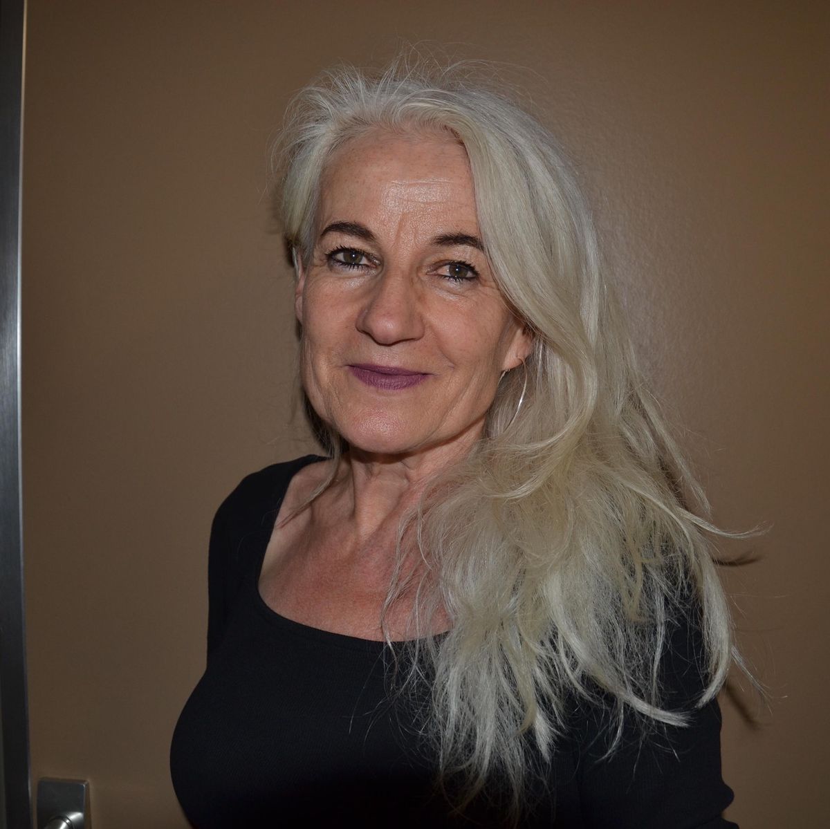 Frau mit langen weissen Haaren und schwarzem Oberteil, stehend vor einer braunen Wand.