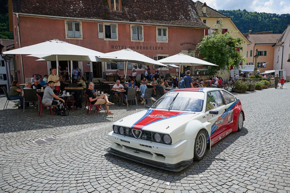 Voiture de course Alfa Romeo garée sur une place pavée devant un café rempli de clients, lors de la course de côte internationale Saint-Ursanne - Les Rangiers, le 17 août 2024.