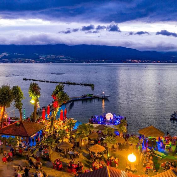 Vue nocturne d’un festival animé au bord du lac Léman, avec des lumières colorées et des stands, en arrière-plan les montagnes.