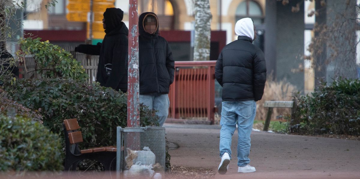 Scène de deal dans le jardin Japonais à côté de la gare d'Yverdon. Deux personnes vêtues de vestes sombres et de capuches blanches se tiennent debout sur un chemin près de bancs et de buissons.