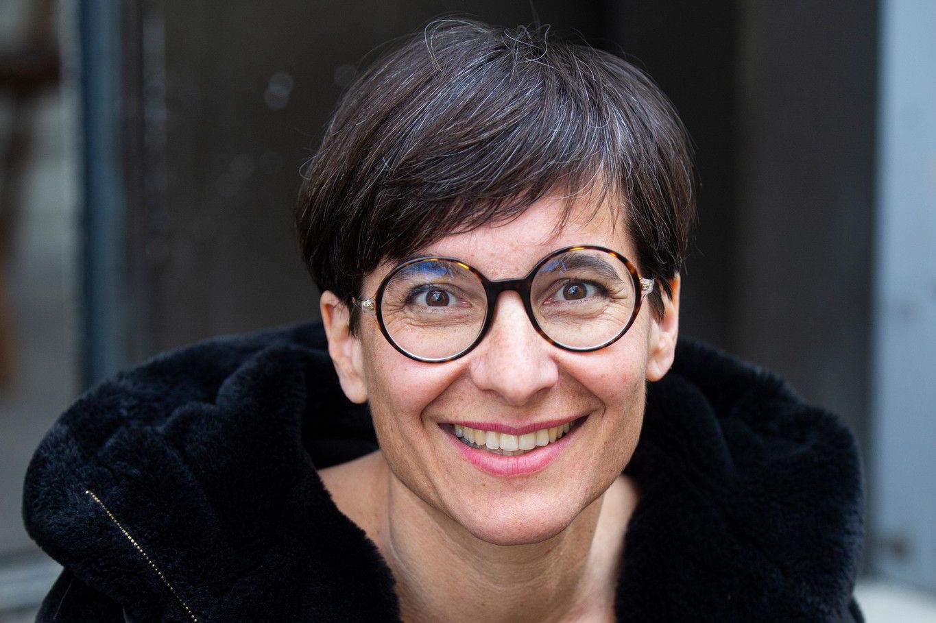 Femme souriante aux cheveux courts, portant des lunettes et un manteau noir, assise dehors.