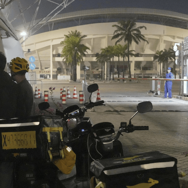 Des livreurs à vélo discutent près d'une route, avec un stade éclairé en arrière-plan et des palmiers visibles de nuit.