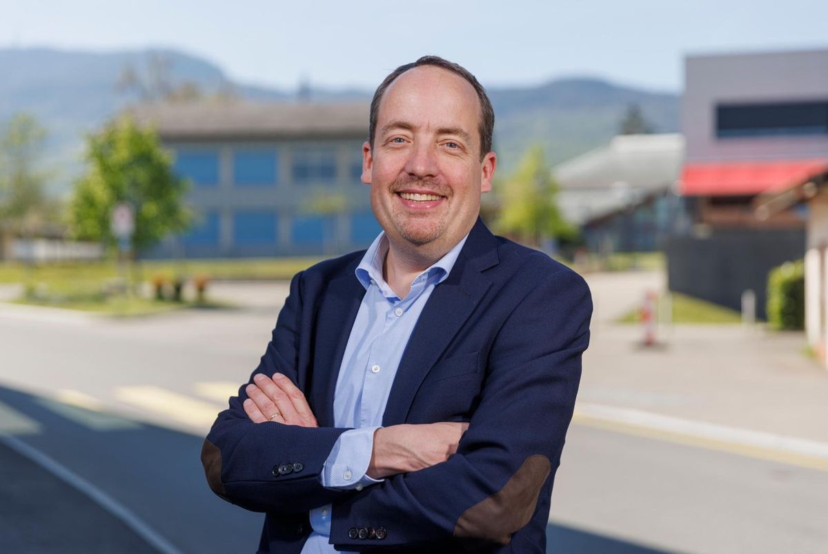 Un homme souriant en costume bleu se tient les bras croisés devant un paysage urbain avec des montagnes en arrière-plan.