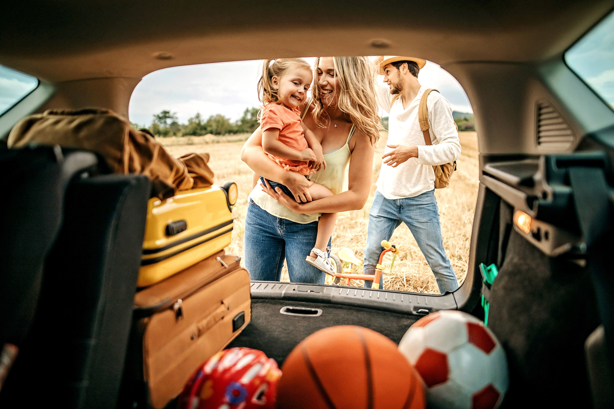 Familie mit Kind belädt Kofferraum für Picknick-Ausflug, mit Koffer, Basketball und Fussball im Vordergrund