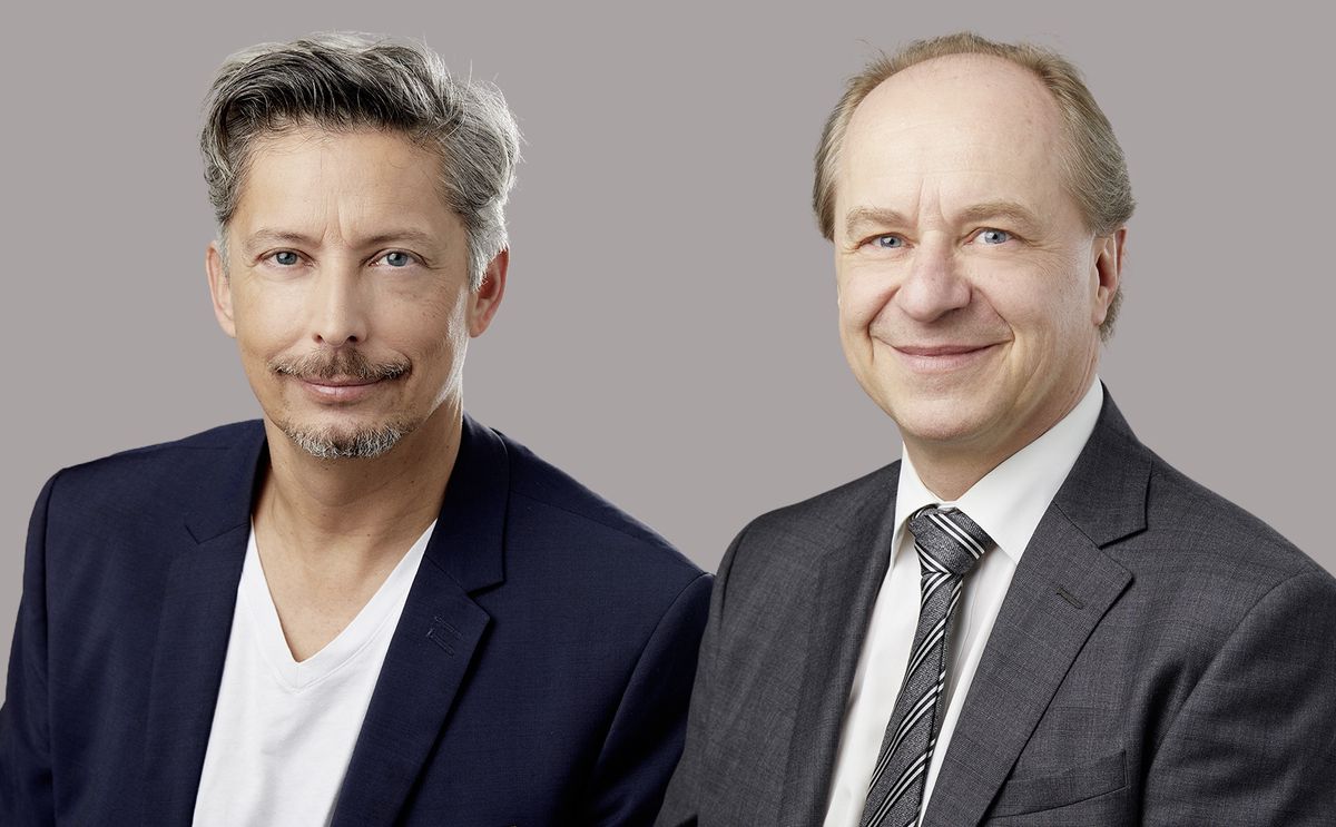 Deux hommes en costume posant pour une photo professionnelle sur un fond gris, l'un portant une veste noire avec un t-shirt blanc et l'autre en costume gris avec une cravate à rayures.