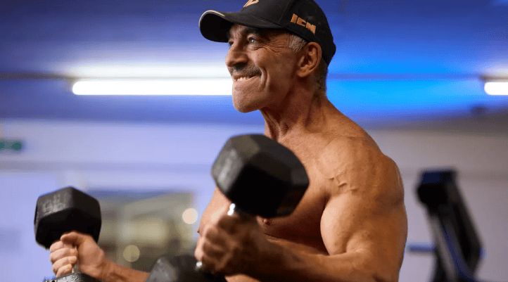 Un homme musclé effectue des exercices de musculation avec des haltères dans une salle de sport, portant une casquette noire.