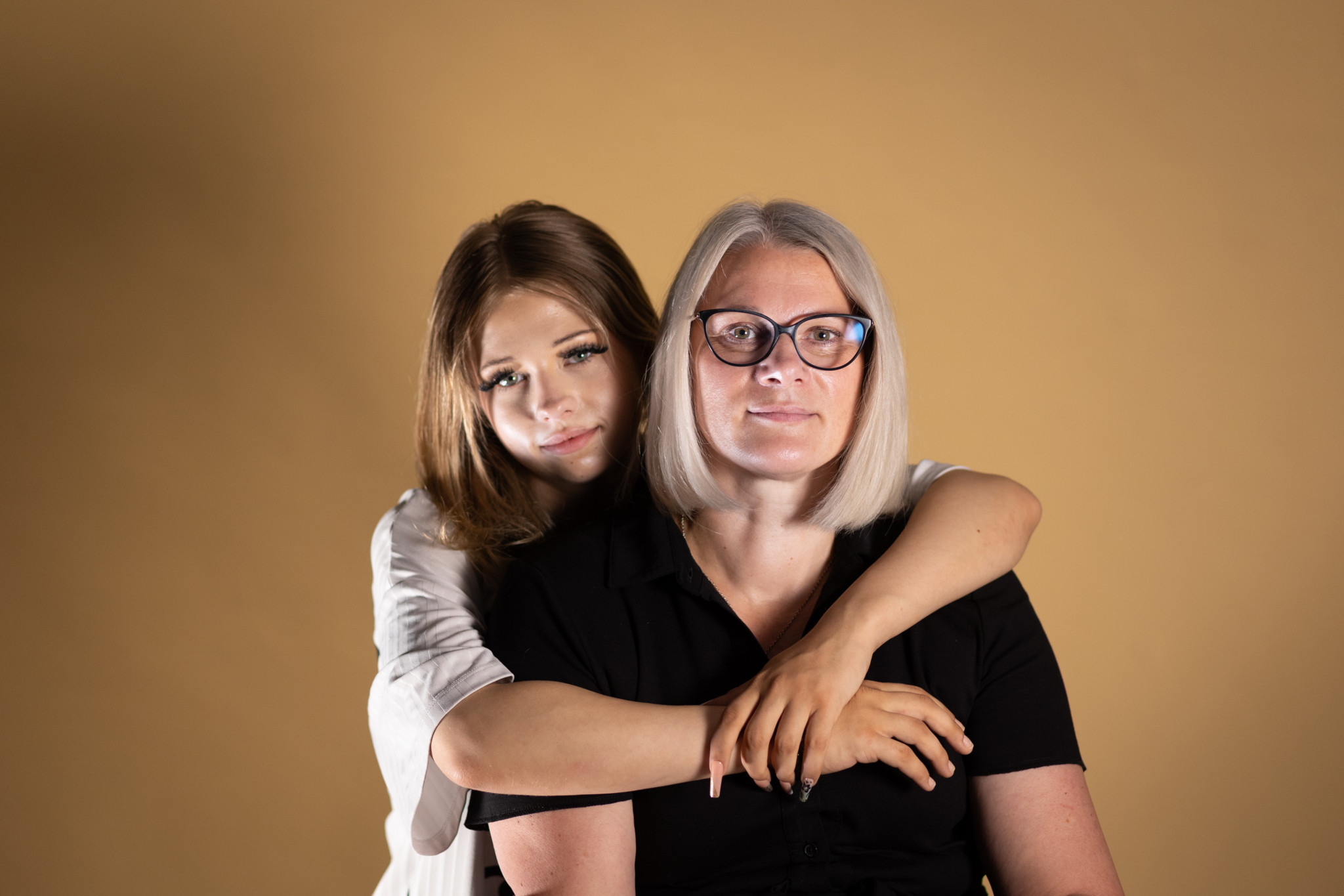Paula L. umarmt ihre Mutter Iwona L. von hinten vor einem beigen Studiohintergrund. Iwona trägt eine Brille und ein schwarzes Shirt.