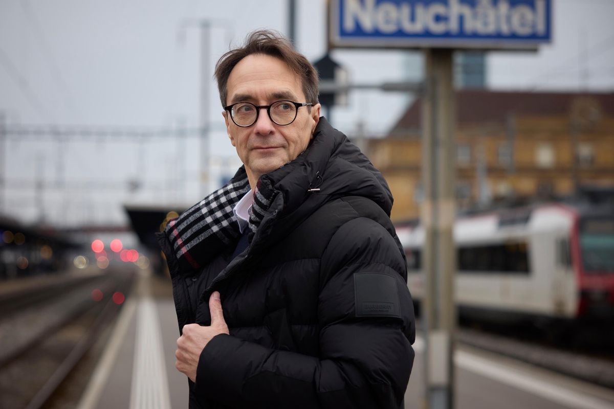 Urs Messerli sur le quai de la gare de Neuchâtel, portant une veste d’hiver, avec un panneau de la gare en arrière-plan.