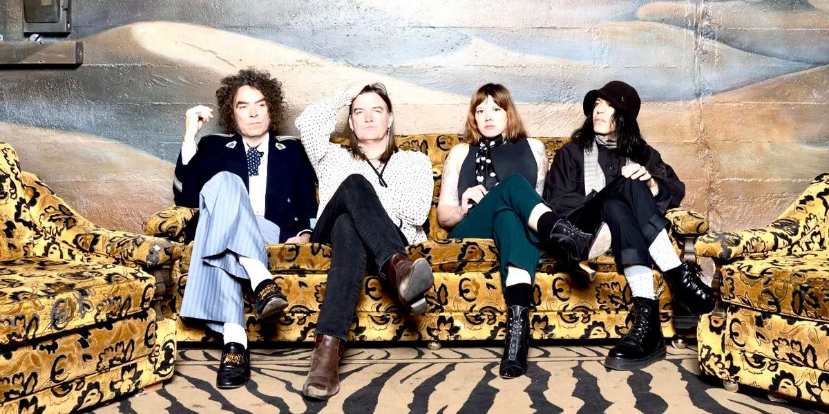 Quatre personnes assises sur un canapé aux motifs floraux, dans une pièce avec une peinture murale de dunes de sable et un ciel nuageux.