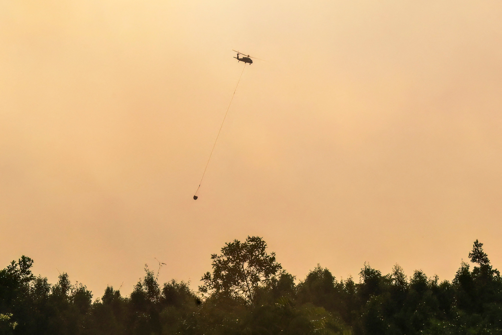 Hubschrauber mit Wasserbehälter bekämpft Torfmoorbrände in Pulang Pisau, Zentralkalimantan, Indonesien, vor orangefarbenem Rauchhimmel.