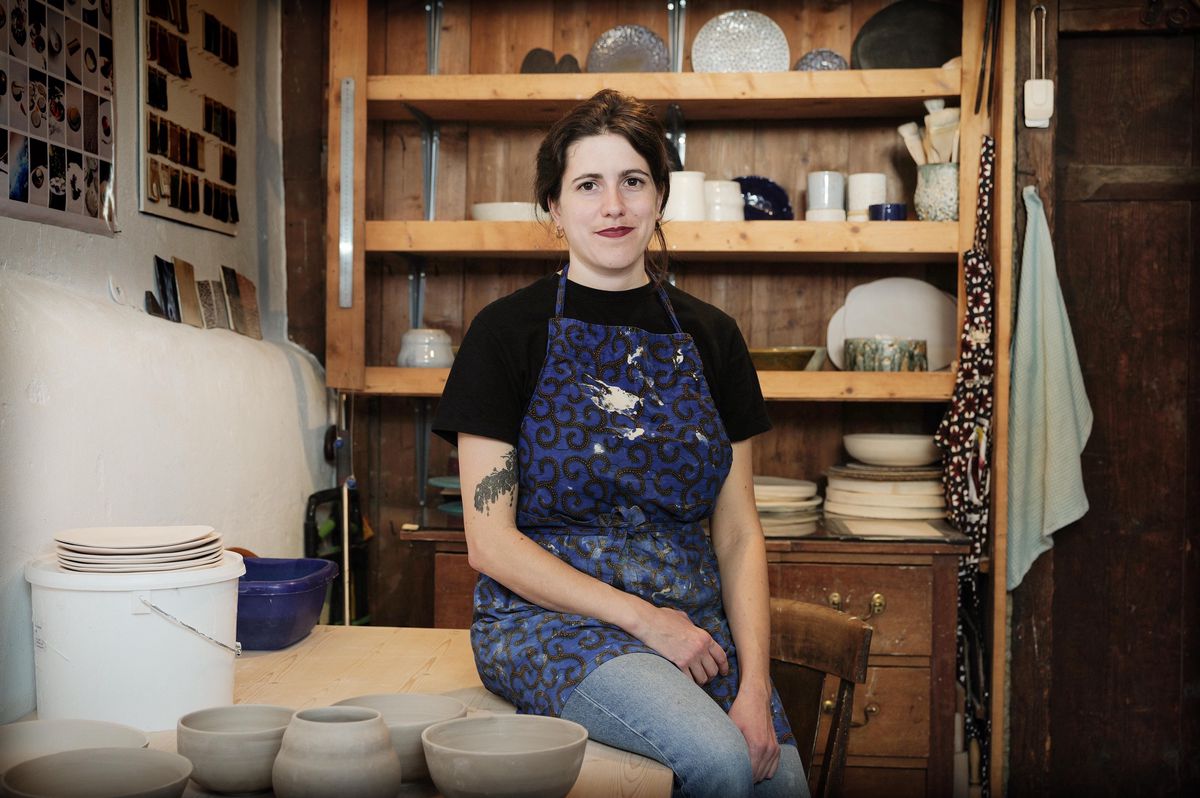 Jeanne Magnenat, jeune céramiste, assise dans son atelier à Genève, entourée de poteries et d'ustensiles de céramique.