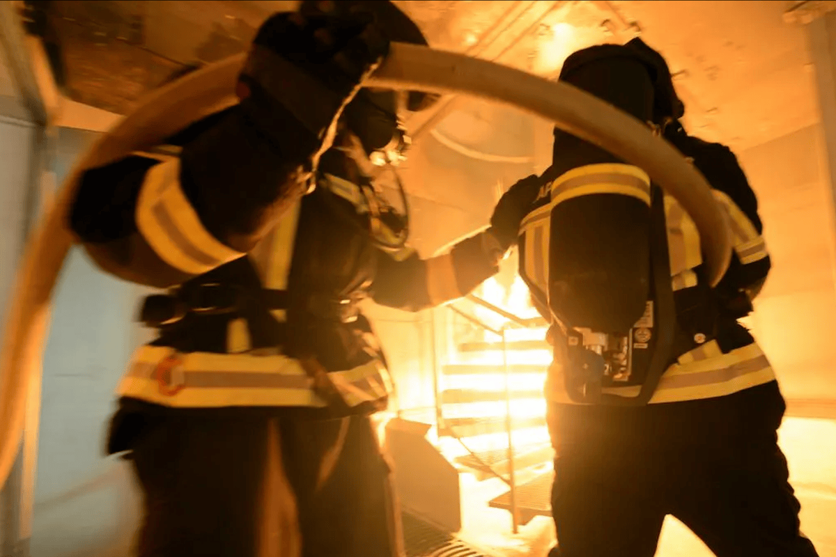 Deux pompiers en tenue de protection tenant un tuyau, combattant un incendie à l’intérieur d’un bâtiment, entourés de fumée et de flammes.