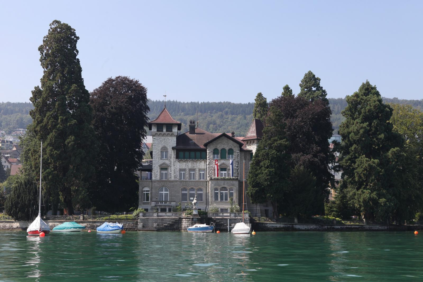 Historische Villa am Seeufer mit mehreren Segelschiffen im Vordergrund, umgeben von hohen Bäumen unter blauem Himmel.