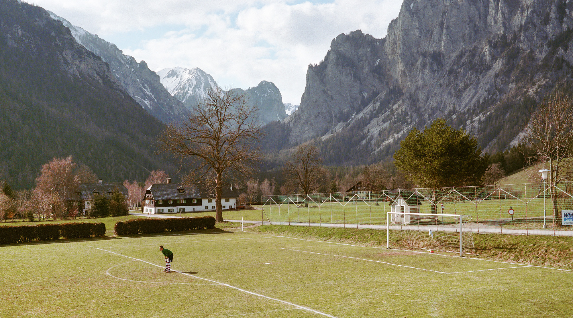Kleines Fussballfeld in alpiner Landschaft mit schneebedeckten Felsgipfeln im Hintergrund und einer einzelnen Person auf dem Rasen.
