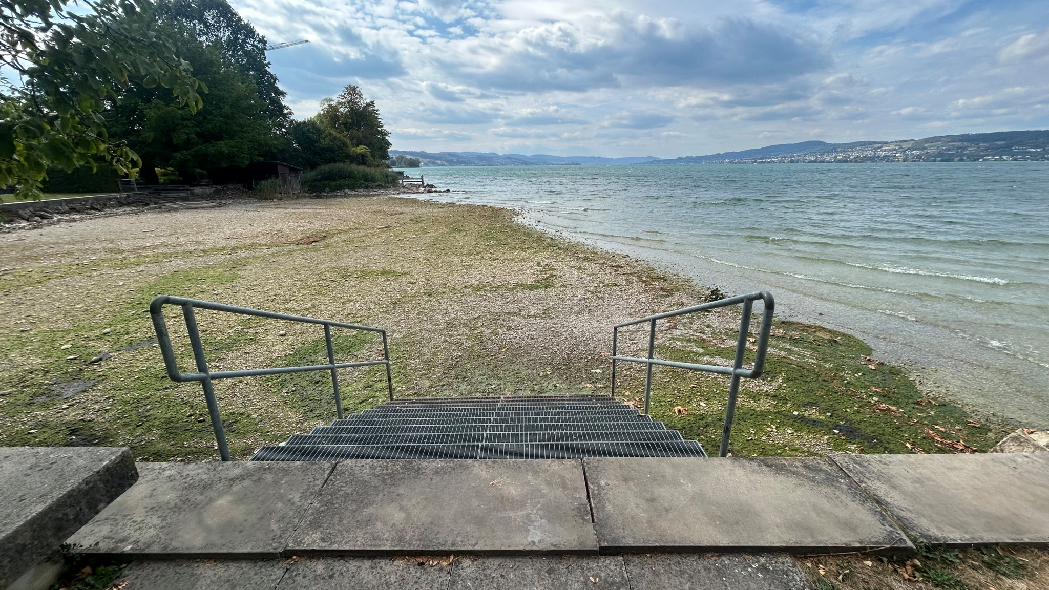Treppe mit Metallgeländer führt zu einem Seeufer mit kiesigem Strand, Bäumen und bewölktem Himmel im Hintergrund.