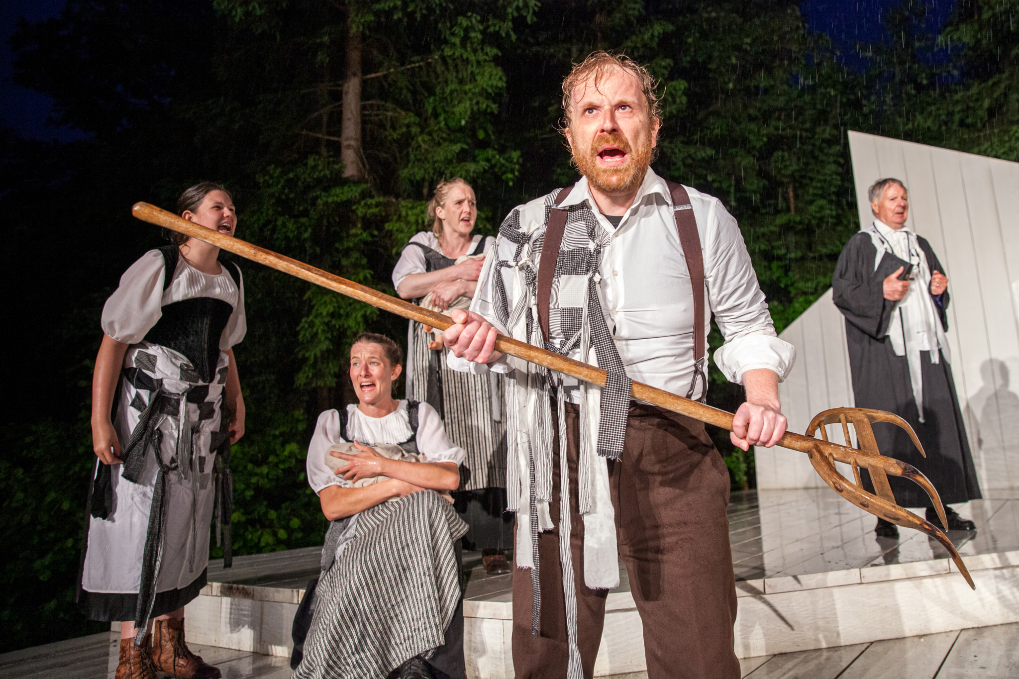 Schauspieler in historischen Kostümen führen ein Theaterstück im Freien auf; ein Mann vorne hält eine Heugabel.