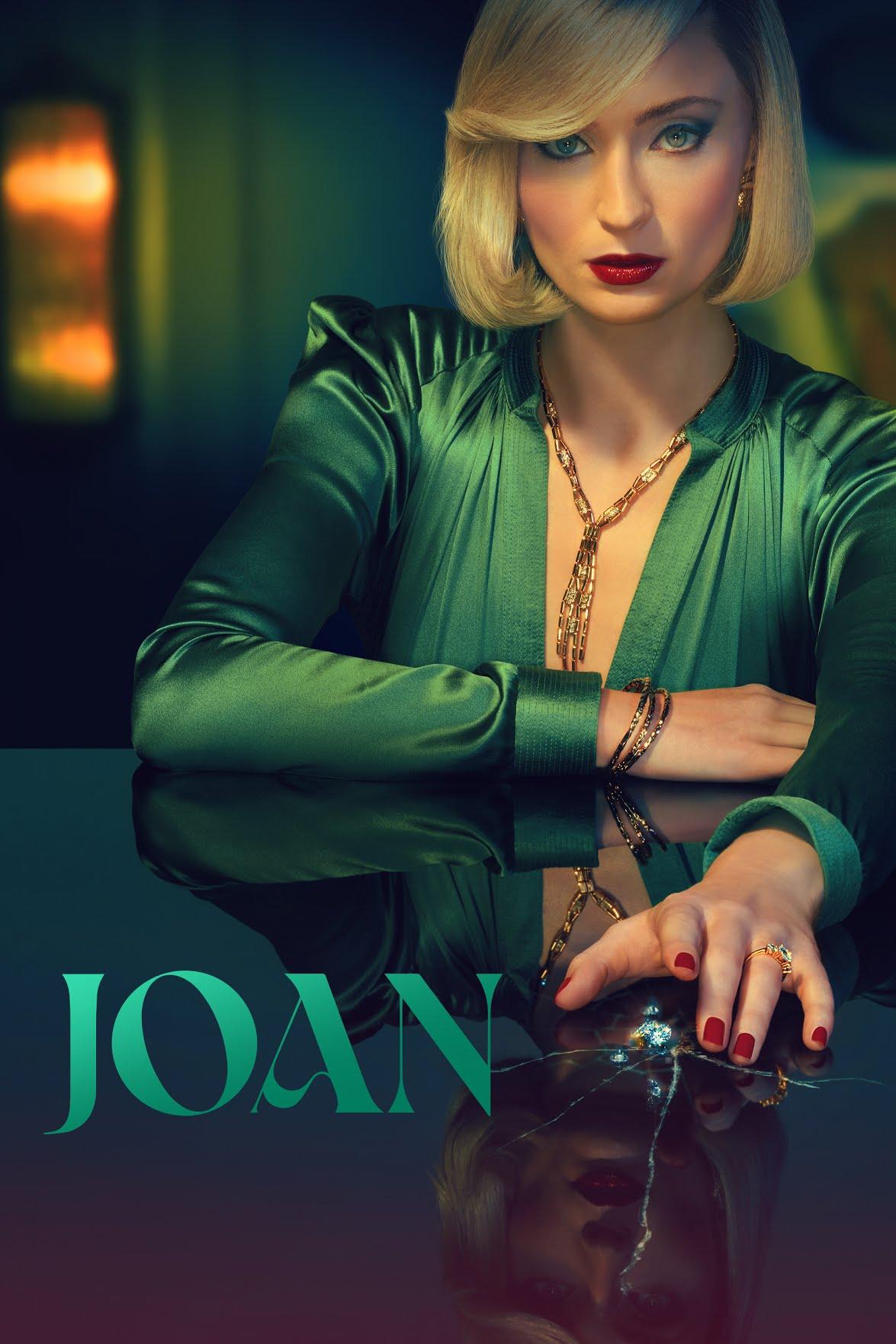 Femme en robe verte élégante avec une coiffure blonde, posant sur une table en verre. Texte affichant ’JOAN’ en lettres vertes stylisées.