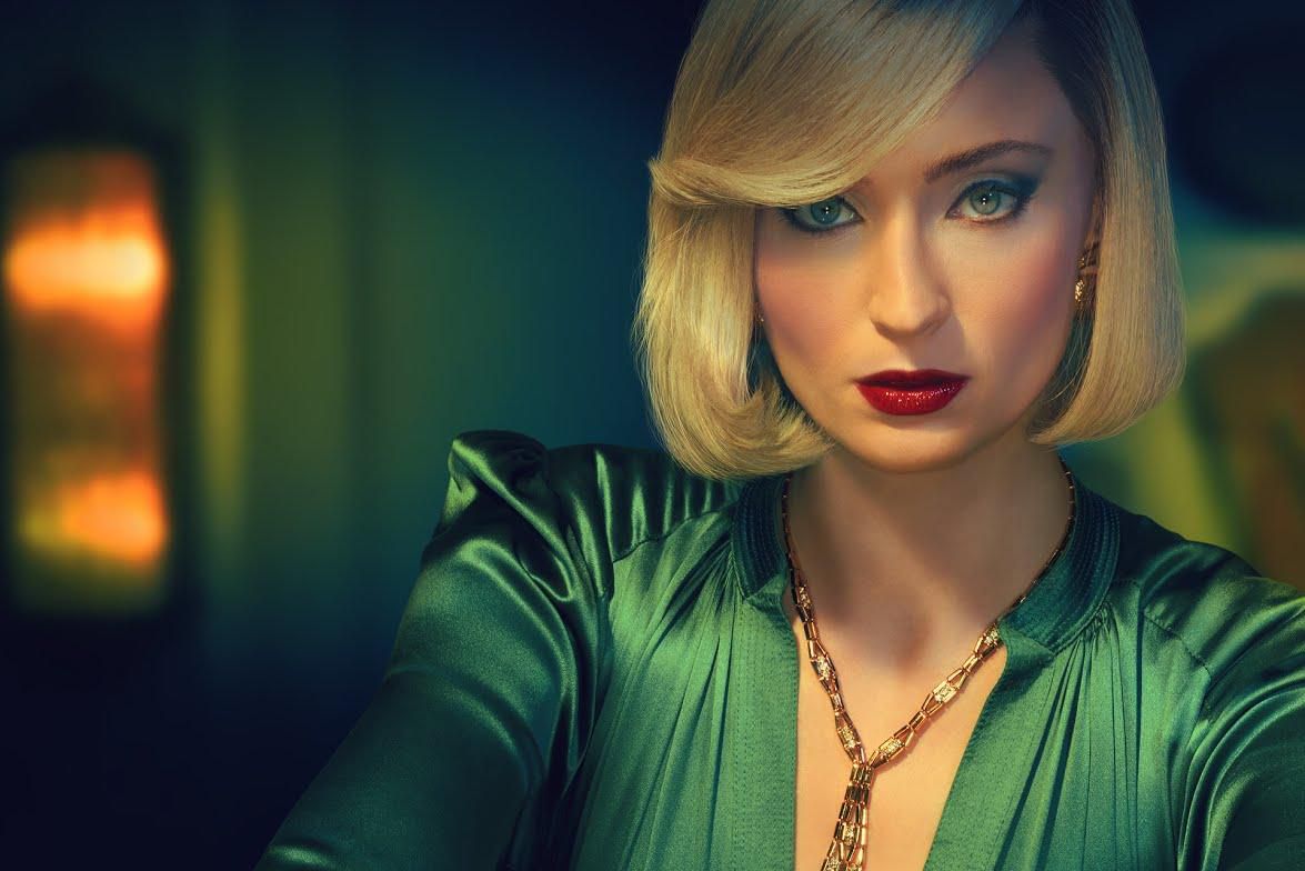 Femme en robe verte élégante avec une coiffure blonde, posant sur une table en verre. Texte affichant ’JOAN’ en lettres vertes stylisées.