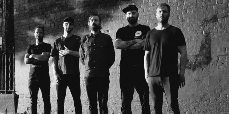 Eine Gruppe von fünf Männern steht nebeneinander vor einer Backsteinmauer, alle in dunkler Kleidung. Sie blicken direkt in die Kamera.