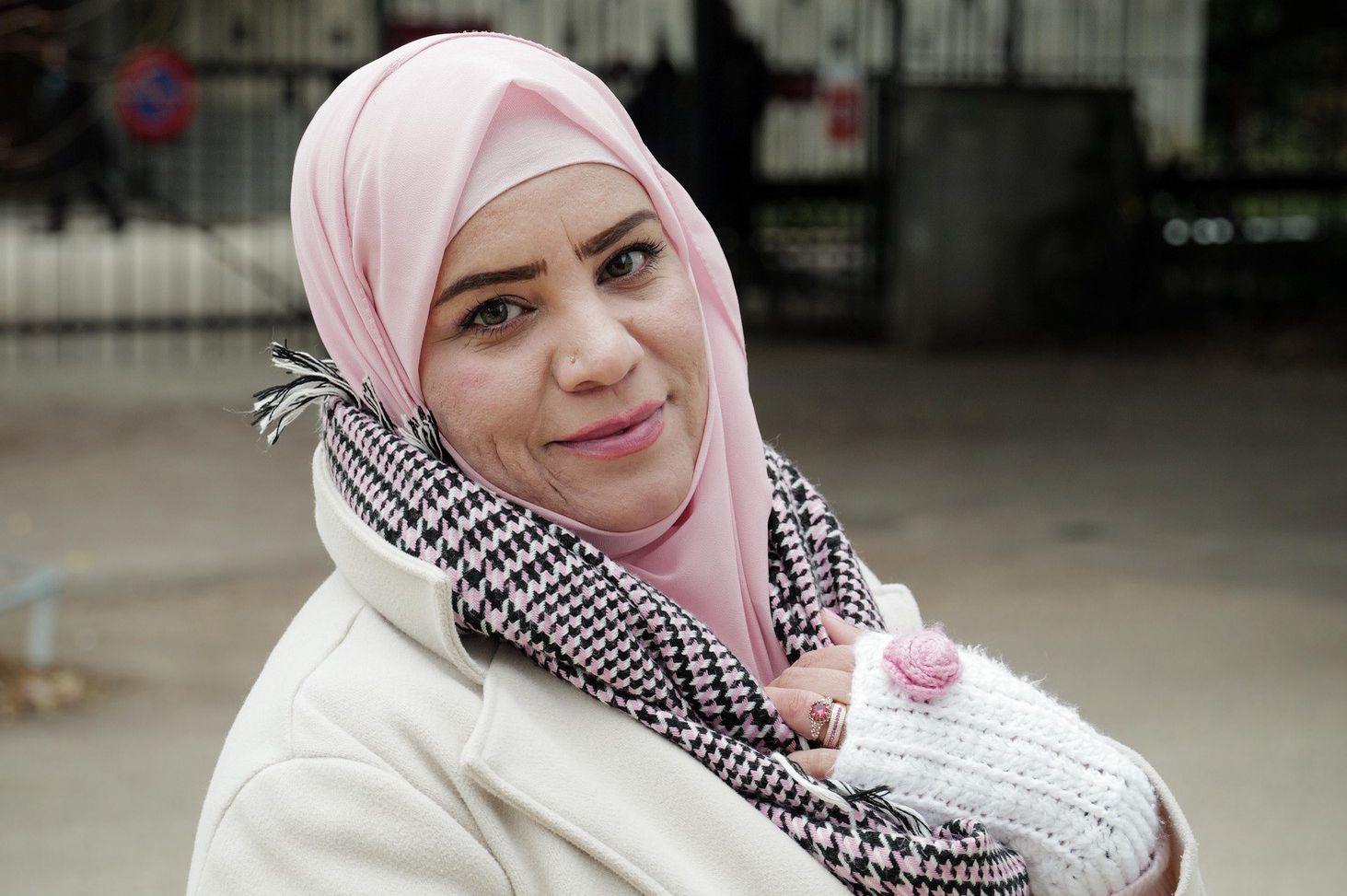 Une femme syrienne à Genève, portant un manteau beige, un foulard à motifs noirs et blancs, et un bonnet rose.
