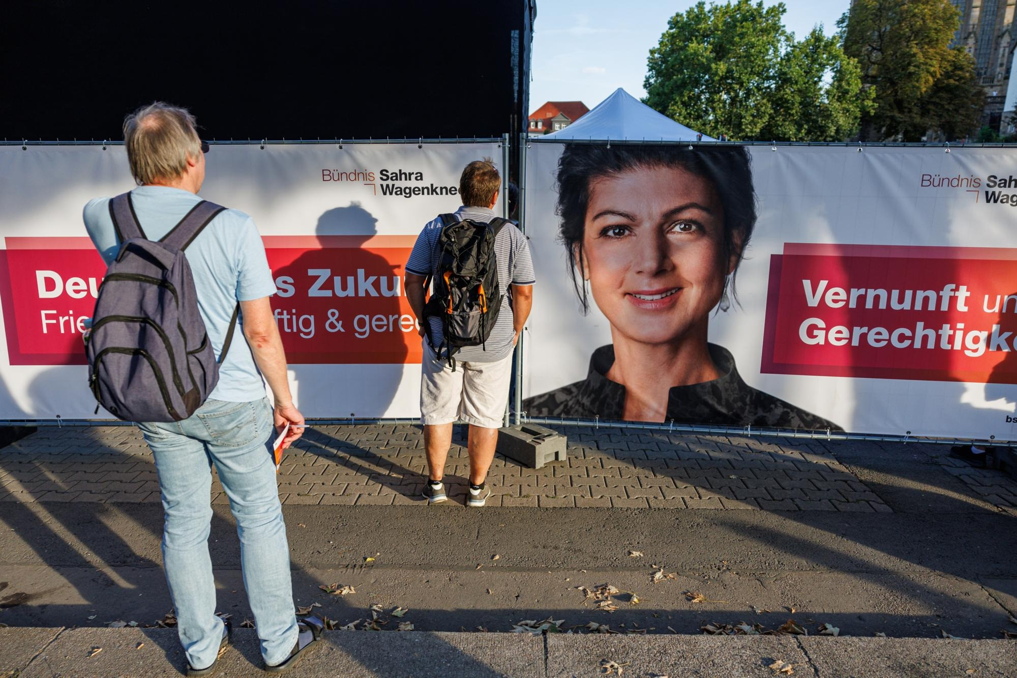 Zwei Personen mit Rucksäcken stehen vor einem Banner mit dem Gesicht von Sahra Wagenknecht und den Worten 'Vernunft und Gerechtigkeit'.
