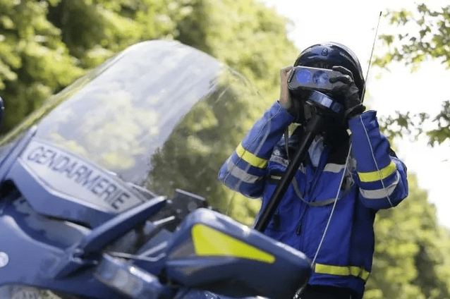 Un gendarme en uniforme bleu utilise un radar de vitesse portable près d'une moto de la gendarmerie, entouré de nature verdoyante.