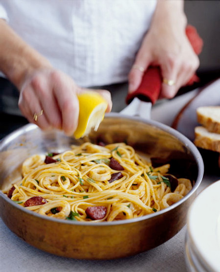 Person presst Zitrone über Spaghetti mit Wurst und Kräutern in einer Kupferpfanne, Brot im Hintergrund.