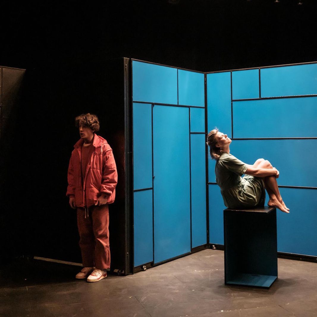 Deux personnes sur scène avec un fond bleu, l’une debout et l’autre assise sur un cube dans un cadre de théâtre, éclairées par des lumières au-dessus.