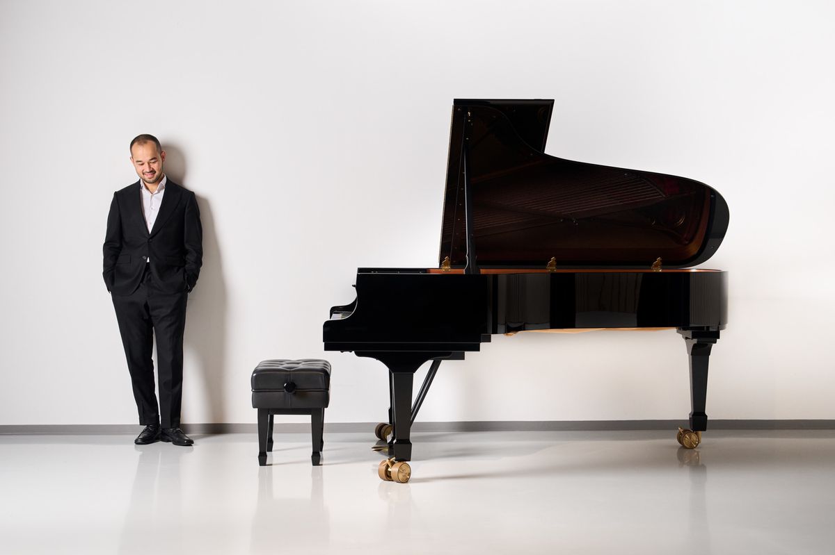 Un homme en costume noir debout à côté d'un piano à queue noir dans une pièce blanche.