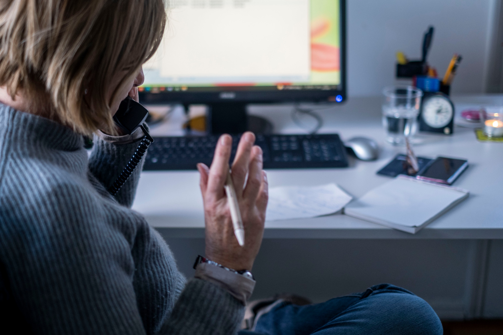 Frau telefoniert in einem Büro, notiert sich etwas mit einem Stift. Auf dem Tisch befinden sich ein Computer, Gläser und ein Notizbuch.