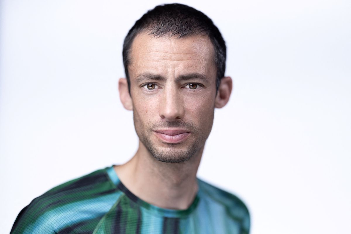 Un homme portant un t-shirt vert et noir regarde directement vers la caméra sur un fond blanc.