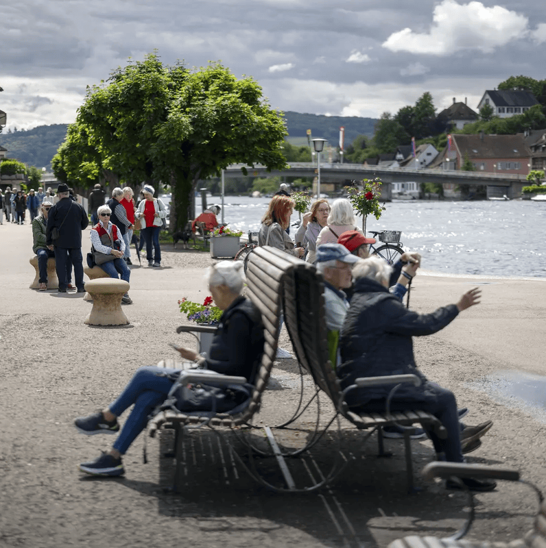 Personnes se détendent sur des bancs le long d’une promenade au bord d’une rivière, avec des bâtiments historiques et un pont en arrière-plan.