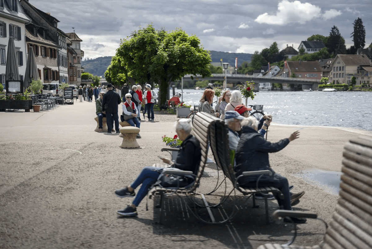 Personnes se détendent sur des bancs le long d’une promenade au bord d’une rivière, avec des bâtiments historiques et un pont en arrière-plan.
