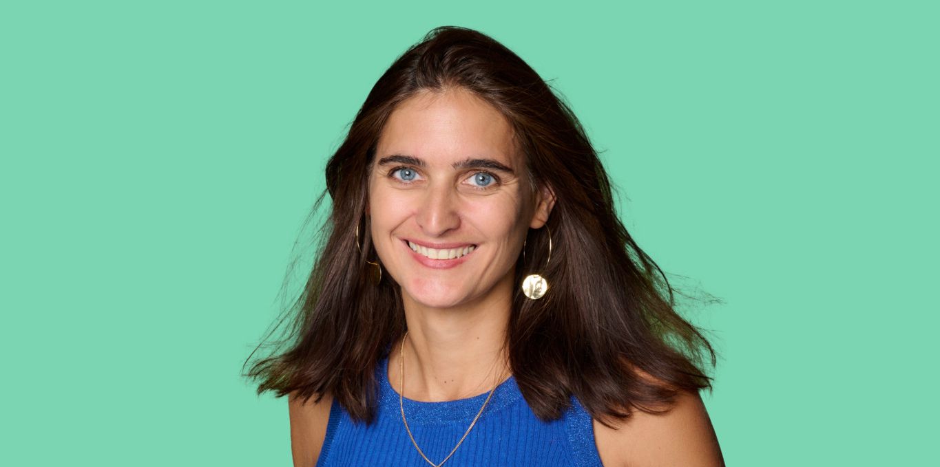 Femme en débardeur bleu et short rouge avec un sourire, mains dans les poches, fond vert.