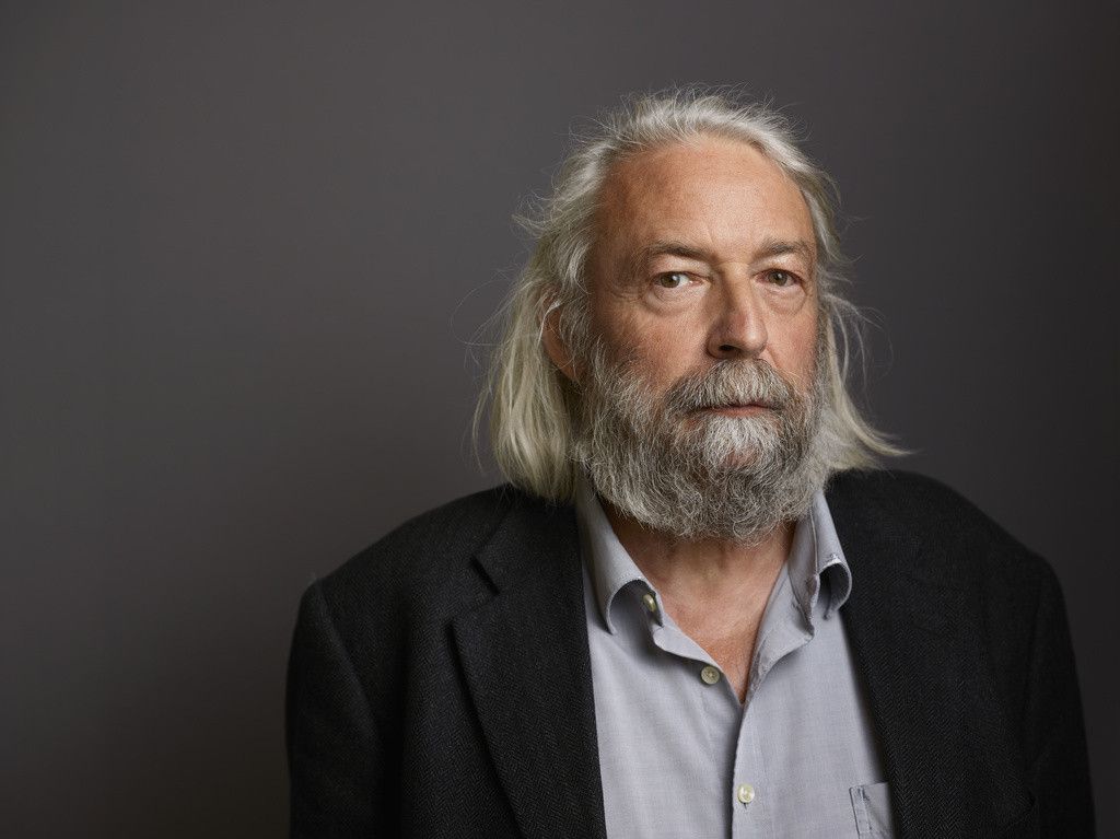 Un homme aux cheveux et à la barbe blanche, portant une veste sombre et une chemise claire, pose devant un fond gris.