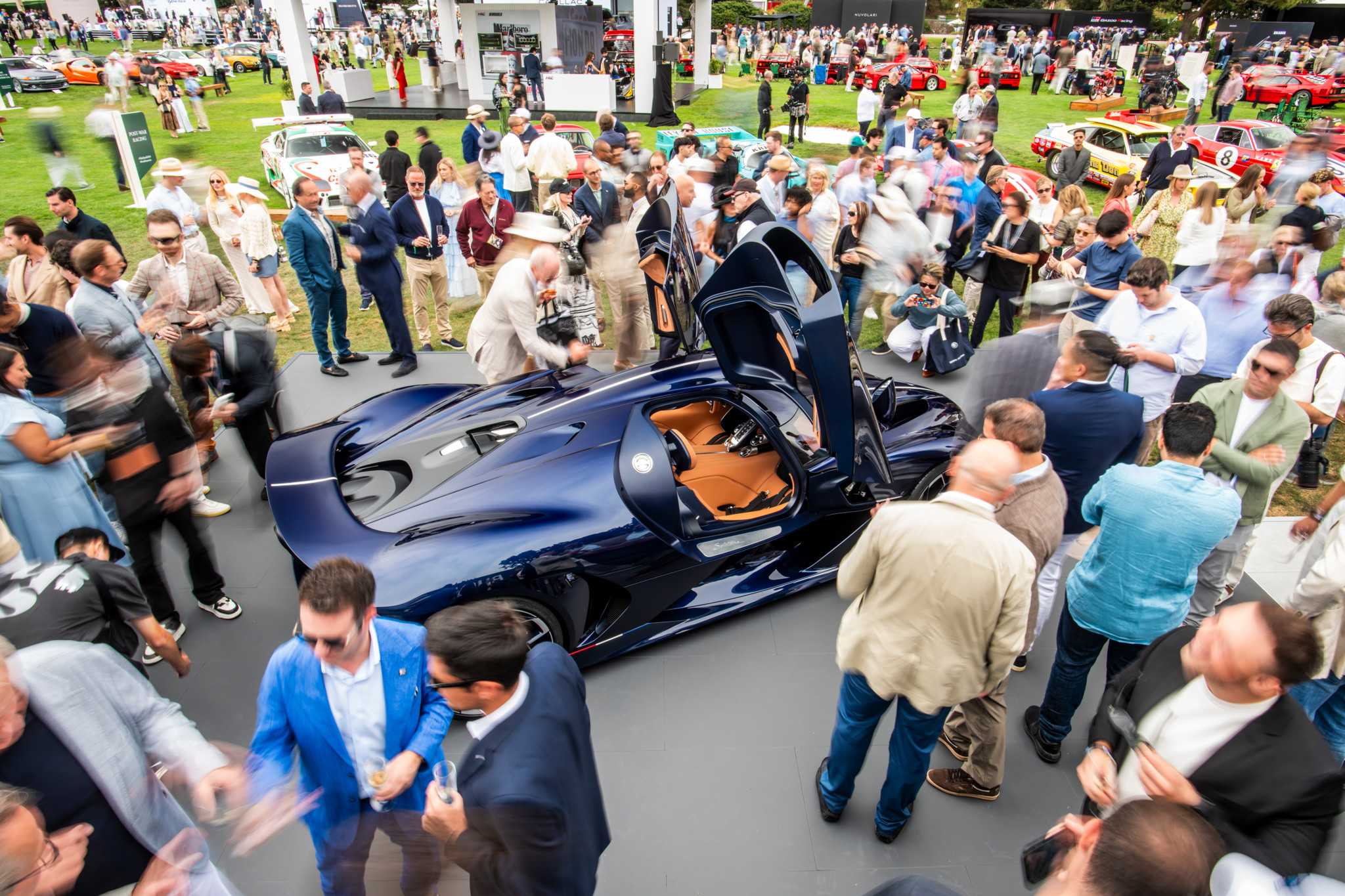 Menschenmenge umringt einen dunkelblauen Supersportwagen mit geöffneten Flügeltüren auf einer Automobilausstellung im Freien.