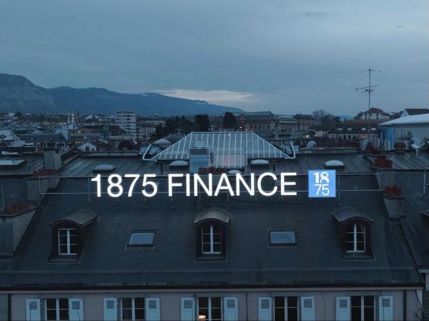 Vue d’un bâtiment avec le logo ’1875 Finance’ illuminé sur le toit, en arrière-plan des montagnes et un ciel nuageux.