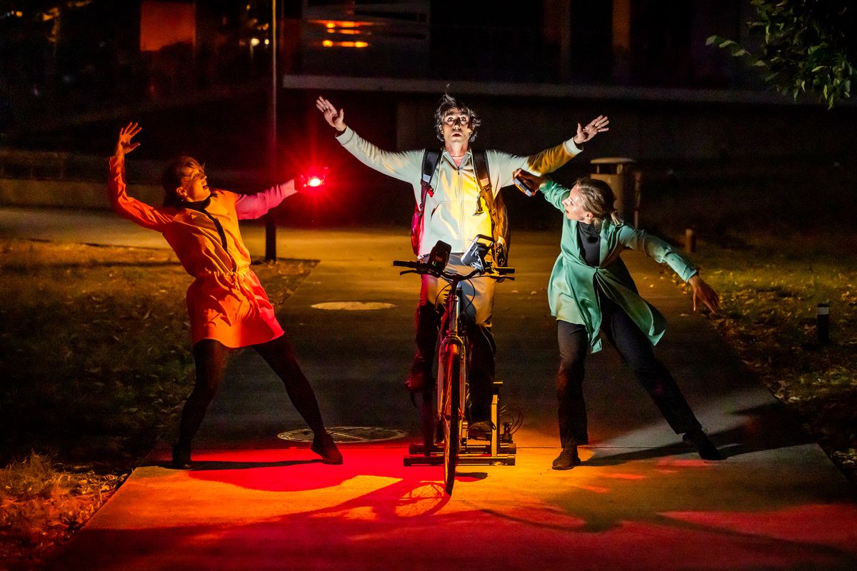 Le spectacle « CUBE » de la Compagnie Zanco au Parc de la Chapelle à Lancy, montre un livreur à vélo entouré de deux danseurs sous un éclairage coloré.
