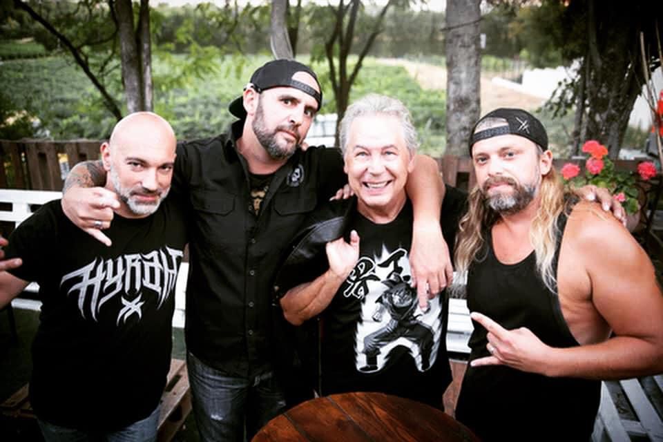 Quatre hommes posant ensemble sur une terrasse, certains portant des vêtements décontractés avec des expressions amicales.