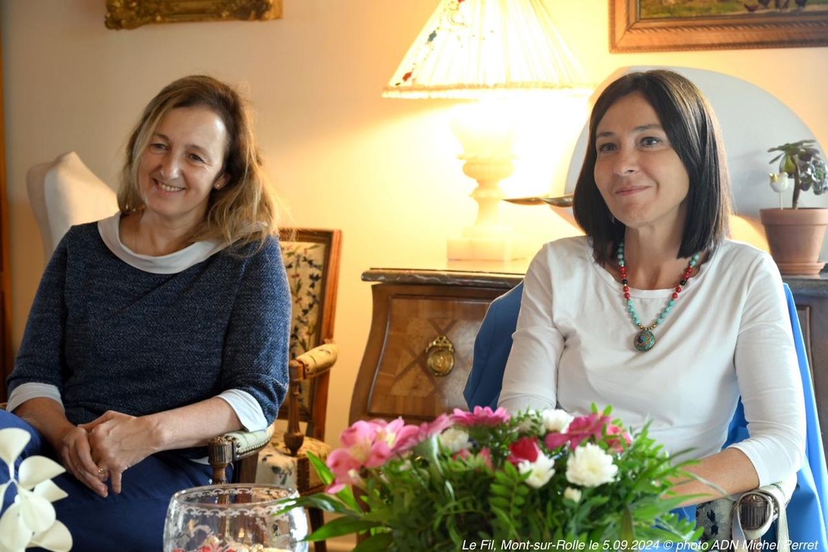 Deux femmes assises dans un salon, souriantes, avec une table garnie de fleurs au premier plan.