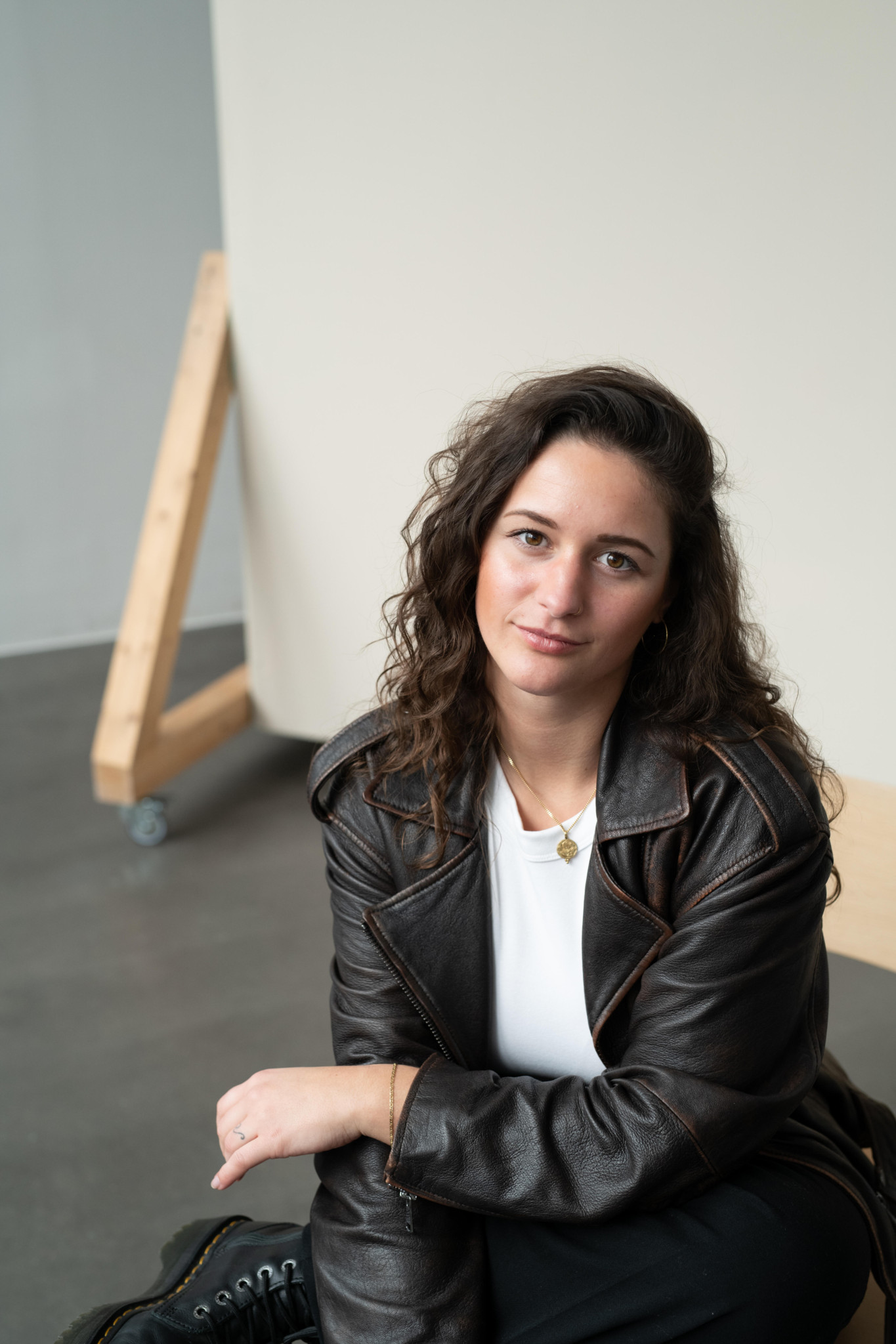 Carolina Wilga mit lockigem braunem Haar und schwarzer Lederjacke sitzt vor einer weissen Wand in einem hellen Raum.
