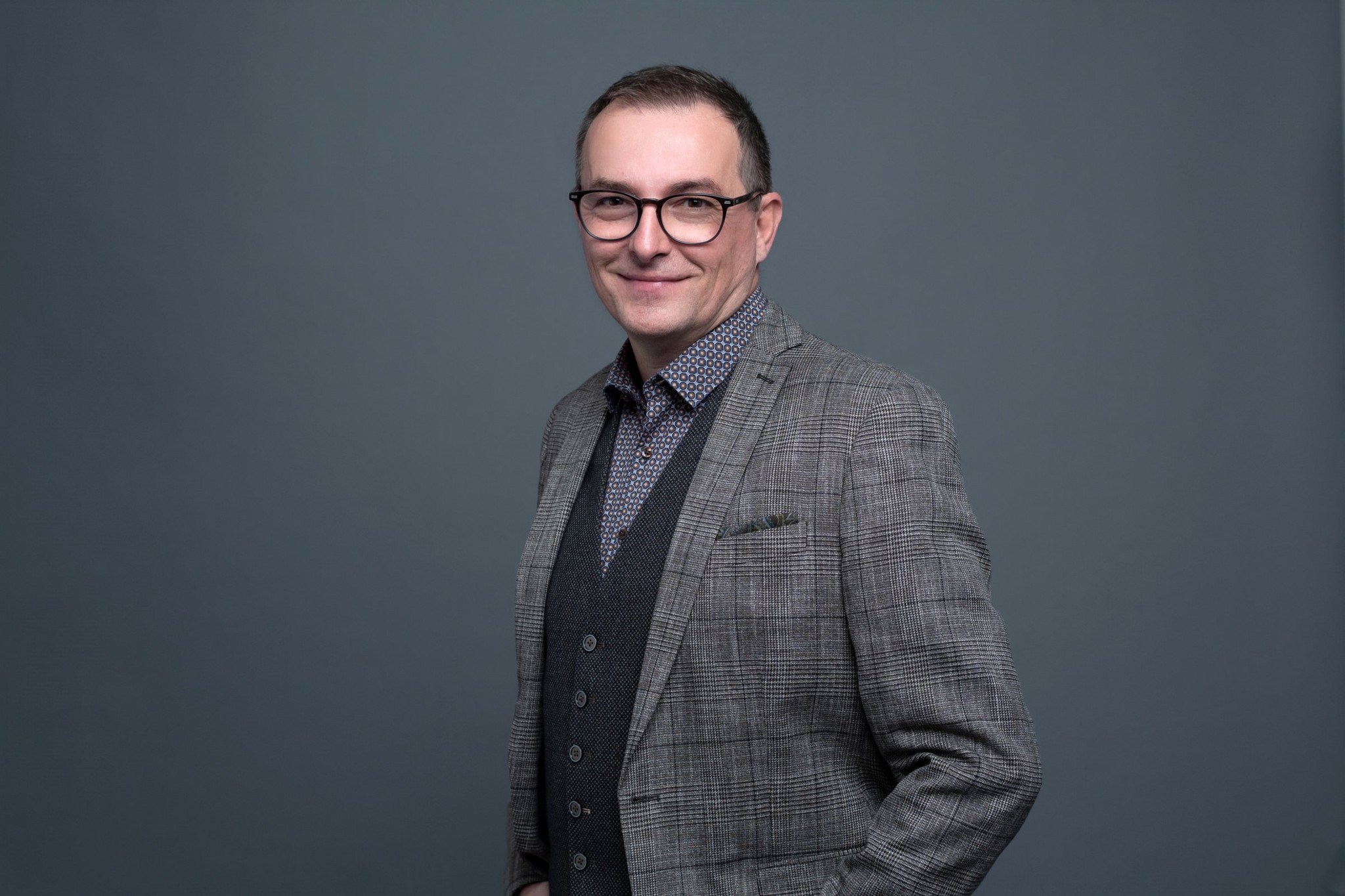 Homme souriant portant des lunettes et un costume gris sur fond gris uni.