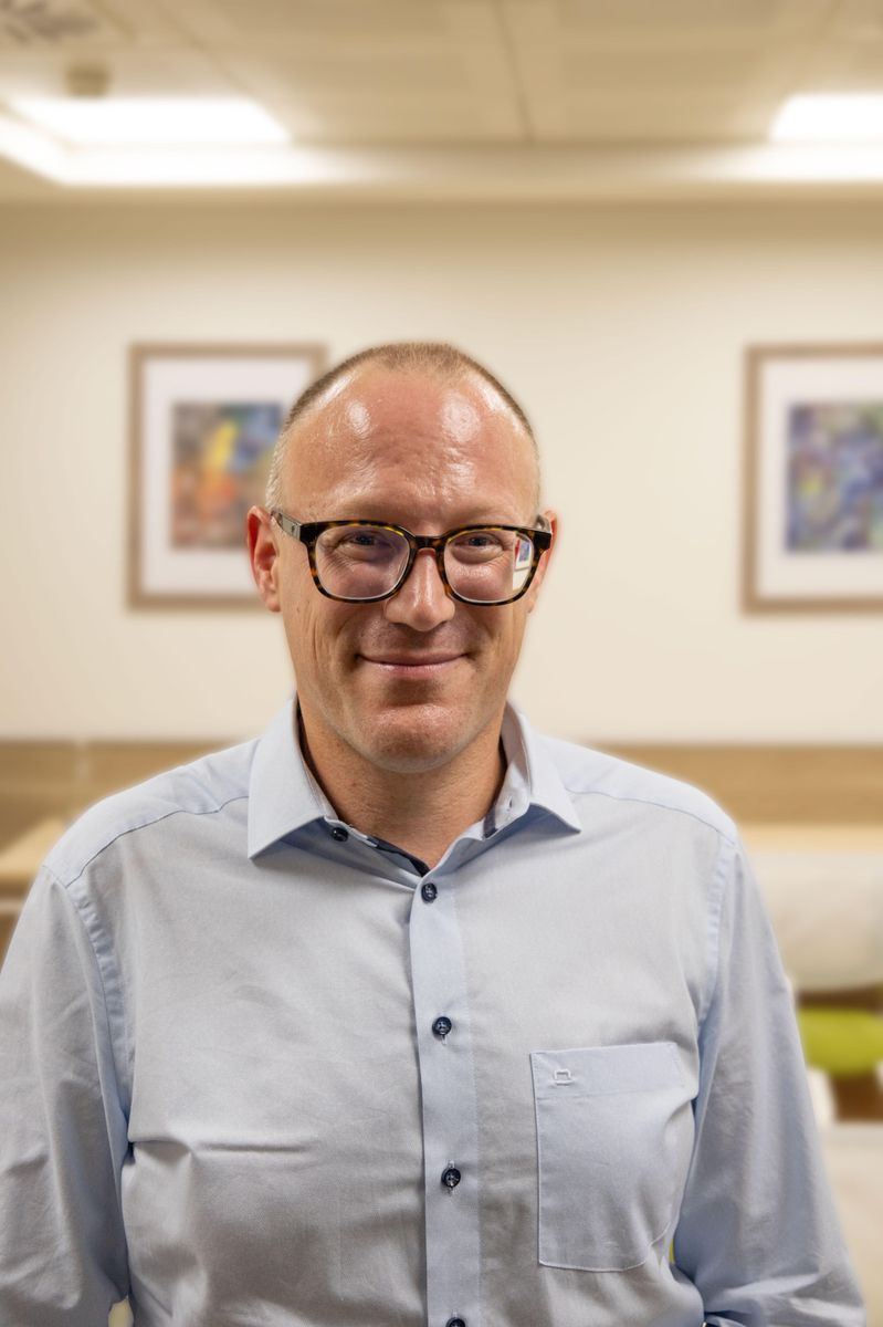 Homme portant des lunettes et une chemise bleue, souriant face à la caméra dans un environnement intérieur avec des tableaux en arrière-plan.