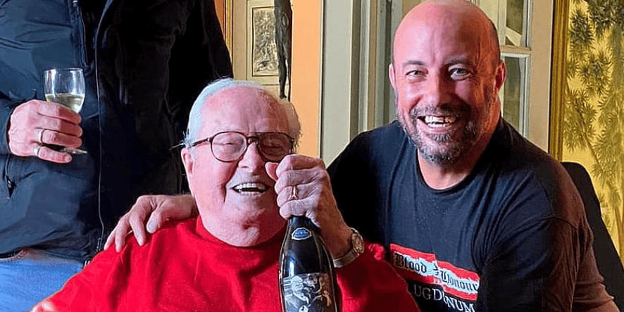 Un homme plus âgé portant un survêtement rouge et des lunettes est assis sur une chaise, souriant, avec une bouteille entre les mains. Un autre homme plus jeune se tient à côté de lui, souriant également, et place sa main sur son épaule.